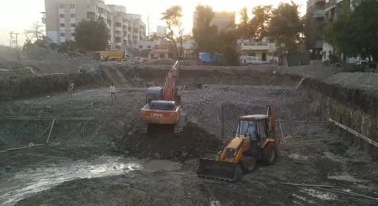 Large building excavation pit with a tracked excavator and a backhoe loader at the bottom, buildings around the site