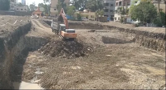 Excavator loading spoil into a tipper truck at the edge of the excavated pit, with buildings beyond