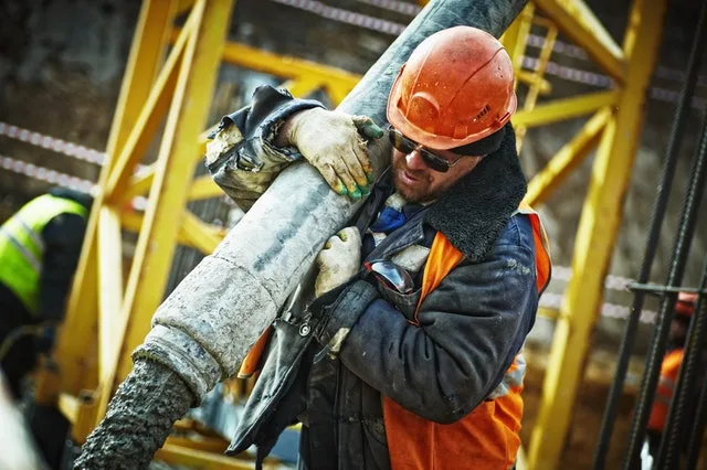 Worker in an orange hard hat and hi-vis vest shouldering a concrete pump hose