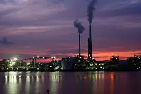 Industrial plant at dusk with two smoking chimneys, its lights reflected in the water