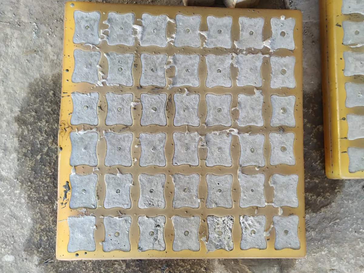 Yellow mould full of set square concrete cover blocks, each with a central wire hole