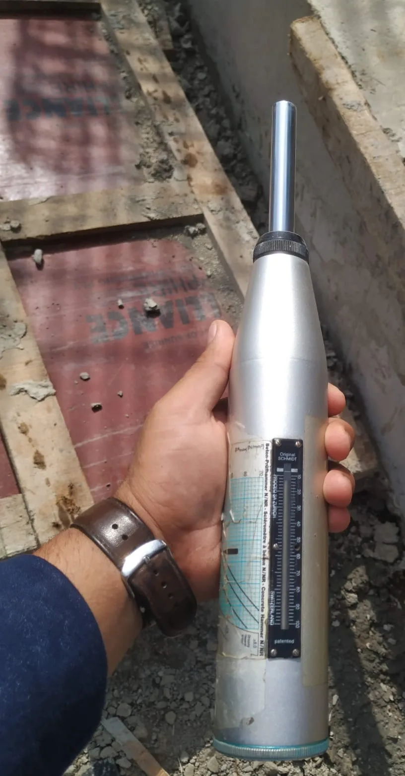 Hand holding a Schmidt rebound hammer upright with the plunger extended, over plywood formwork on site