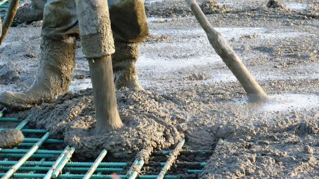 Worker in rubber boots placing wet concrete from a hose onto a mesh of green-coated rebar