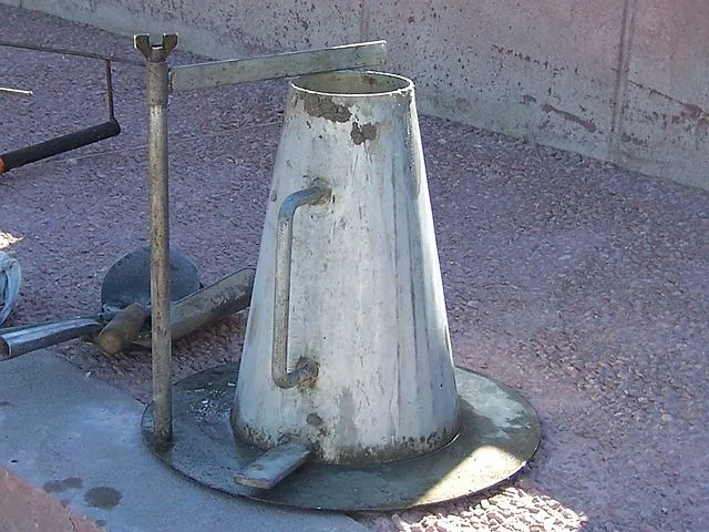 Steel slump cone with side handle and foot piece on a round base plate, with an upright rod and cross-arm beside it