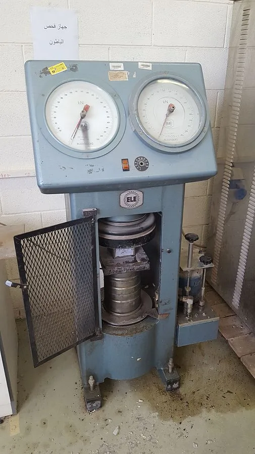Compression testing machine with two dial gauges, its mesh guard door open to show a specimen between the platens