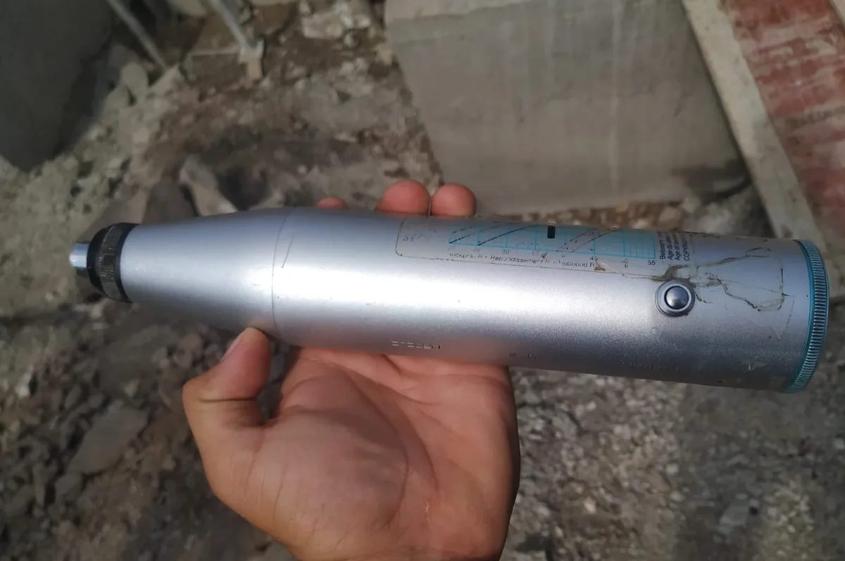 Hand holding a silver Schmidt rebound hammer horizontally over rubble on a construction site
