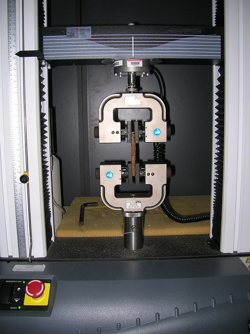 Universal testing machine with a flat specimen clamped between upper and lower grips, emergency stop at the base